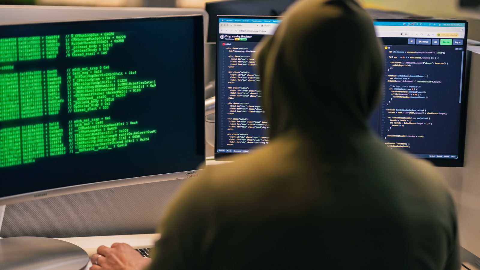 Person in a hooded top seen from behind, working at two monitors showing code