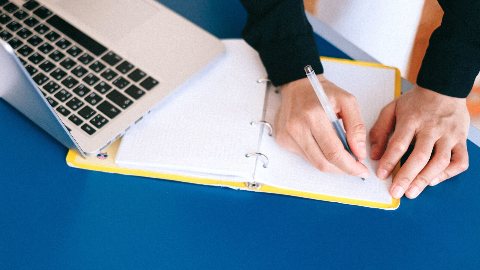 Hands writing in a ring notebook on a blue desk next to an open laptop